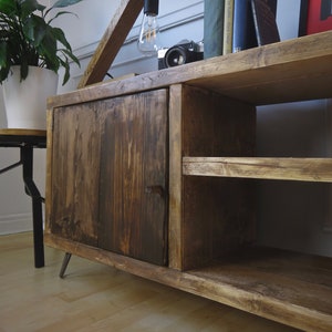 K&ouml;nnte beinhalten: Rustikaler Holzschrank mit dunkel gebeizter Oberfl&auml;che. Der Schrank hat eine T&uuml;r, ein offenes Regal und Metallbeine. Eine Holzlampe mit Gl&uuml;hbirne, B&uuml;cher und eine Kamera stehen obenauf. Links ist eine Topfpflanze zu sehen. Der Schrank ist ca. 120 cm breit.
