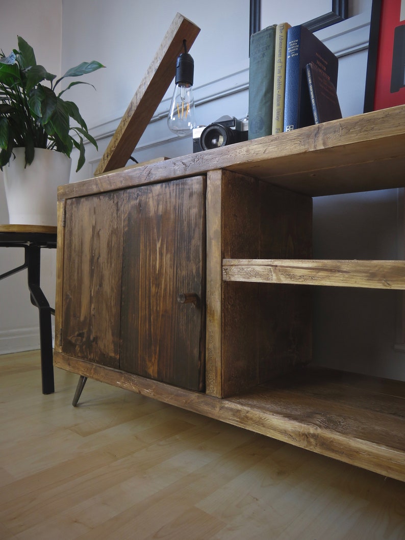 K&ouml;nnte beinhalten: Rustikaler Holzschrank mit dunkel gebeizter Oberfl&auml;che. Der Schrank hat eine T&uuml;r, ein offenes Regal und Metallbeine. Eine Holzlampe mit Gl&uuml;hbirne, B&uuml;cher und eine Kamera stehen obenauf. Links ist eine Topfpflanze zu sehen. Der Schrank ist ca. 120 cm breit.