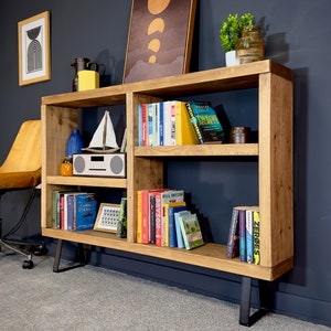 May include: A wooden bookshelf with multiple shelves, filled with books of various colors. The bookshelf has a natural wood finish and black metal legs. Decorative items like a sailboat model and a small radio are also visible.
