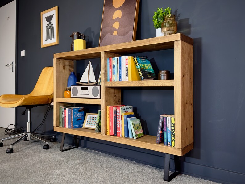May include: A wooden bookshelf with multiple shelves, filled with books of various colors. The bookshelf has a natural wood finish and black metal legs. Decorative items like a sailboat model and a small radio are also visible.