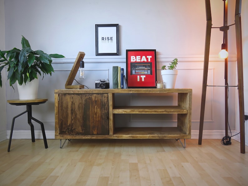 K&ouml;nnte beinhalten: Rustikaler TV-Schrank aus Holz mit T&uuml;r und offenen Regalen. Ein gerahmter Druck mit dem Text "BEAT IT" h&auml;ngt dar&uuml;ber. Eine Holzlampe mit Gl&uuml;hbirne und eine Vintage-Kamera stehen darauf. Eine Stehlampe und Topfpflanzen sind ebenfalls im Raum. Der Schrank ist etwa 120 cm breit.