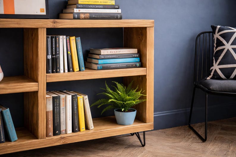 Op de afbeelding: Houten boekenkast met meerdere planken gevuld met boeken van verschillende formaten en kleuren. Een kleine potvaren in een blauwe pot staat op een plank. De boekenkast heeft zwarte metalen haarspeldpoten.