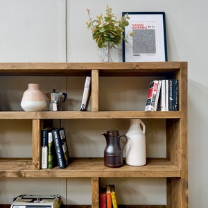 Puede incluir: Una estanter&iacute;a r&uacute;stica de madera llena de libros, jarrones decorativos, una cafetera y una impresi&oacute;n enmarcada. La estanter&iacute;a tiene varios estantes y est&aacute; te&ntilde;ida de un color marr&oacute;n c&aacute;lido. La impresi&oacute;n presenta el texto "YAYOI KUSAMA".
