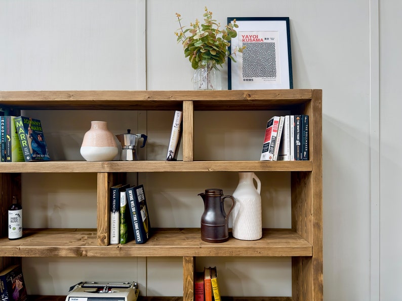 Puede incluir: Una estanter&iacute;a r&uacute;stica de madera llena de libros, jarrones decorativos, una cafetera y una impresi&oacute;n enmarcada. La estanter&iacute;a tiene varios estantes y est&aacute; te&ntilde;ida de un color marr&oacute;n c&aacute;lido. La impresi&oacute;n presenta el texto "YAYOI KUSAMA".