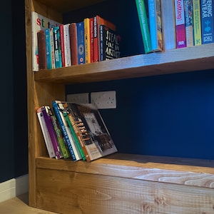May include: A wooden bookshelf with multiple shelves filled with books. The shelves are stained a light brown color. The background wall is a dark blue color. The books are a variety of colors and sizes. Some of the book titles are visible, including "Steal Like an Artist", "Modern Rustic", and "Gardening Through the Year".