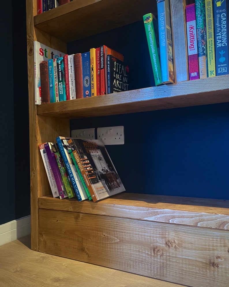 May include: A wooden bookshelf with multiple shelves filled with books. The shelves are stained a light brown color. The background wall is a dark blue color. The books are a variety of colors and sizes. Some of the book titles are visible, including "Steal Like an Artist", "Modern Rustic", and "Gardening Through the Year".