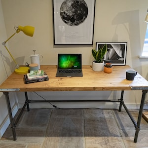 May include: A rustic wooden desk with black metal pipe legs. The desk has a laptop, books, a yellow lamp, a plant, and a mug on top. The desk is in a room with white walls and a window.