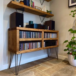 May include: A rustic wooden media console with hairpin legs, filled with CDs. Two speakers and audio components sit on top, with a matching shelf above. A potted plant with green leaves is to the right.
