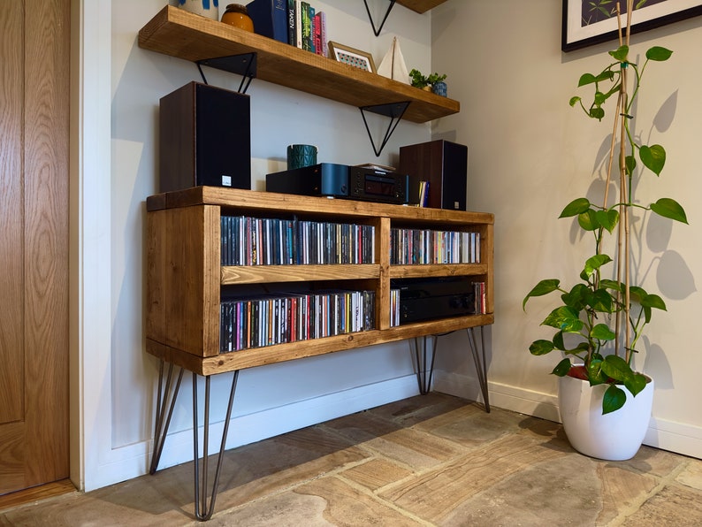 May include: A rustic wooden media console with hairpin legs, filled with CDs. Two speakers and audio components sit on top, with a matching shelf above. A potted plant with green leaves is to the right.