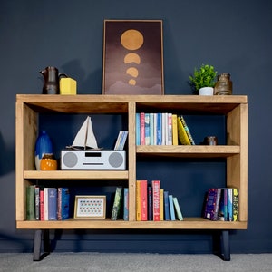 May include: A wooden bookshelf with multiple shelves, displaying books, decorative items, and a framed print. The bookshelf has a natural wood finish, black legs, and is set against a dark blue wall. Various items are arranged on the shelves.