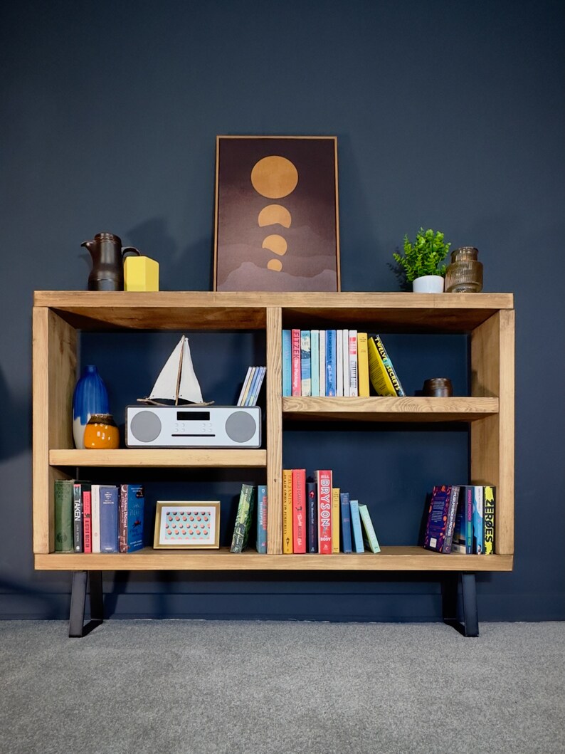 May include: A wooden bookshelf with multiple shelves, displaying books, decorative items, and a framed print. The bookshelf has a natural wood finish, black legs, and is set against a dark blue wall. Various items are arranged on the shelves.
