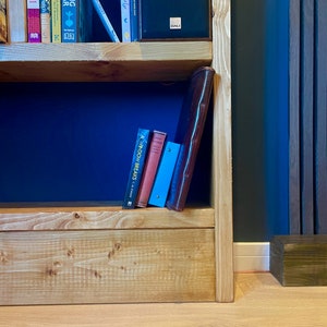 May include: A wooden bookshelf with two shelves, filled with books and a brown leather binder. The bookshelf is against a dark blue wall with vertical wood paneling.