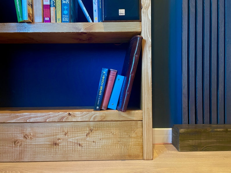 May include: A wooden bookshelf with two shelves, filled with books and a brown leather binder. The bookshelf is against a dark blue wall with vertical wood paneling.