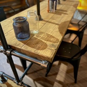 K&ouml;nnte beinhalten: Rustikaler Holztisch mit Metallrohrrahmen, mit einer blauen Glasvase und einem klaren Glasbeh&auml;lter. Schwarze Metallst&uuml;hle sind um den Tisch positioniert. Der Tisch ist etwa 152 cm lang.