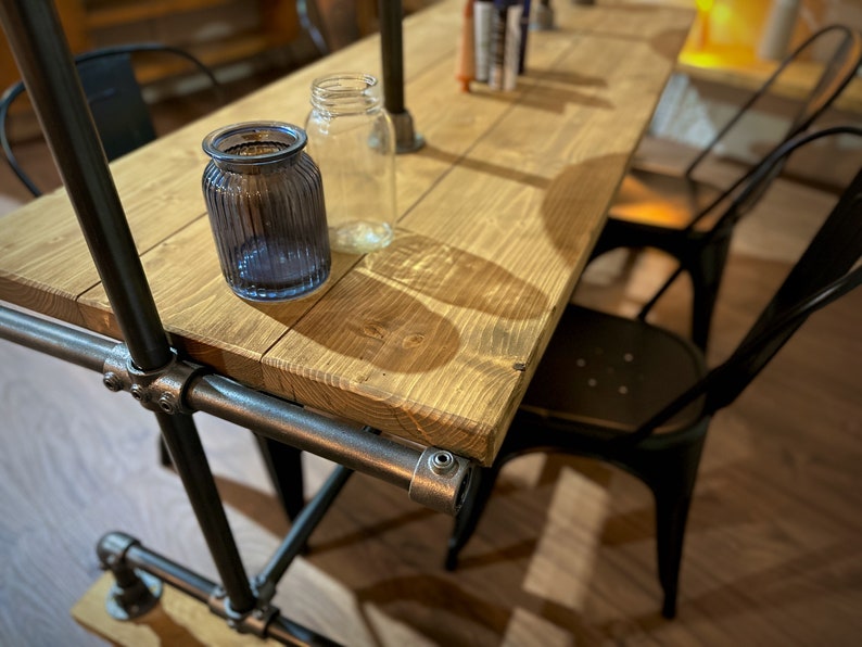 K&ouml;nnte beinhalten: Rustikaler Holztisch mit Metallrohrrahmen, mit einer blauen Glasvase und einem klaren Glasbeh&auml;lter. Schwarze Metallst&uuml;hle sind um den Tisch positioniert. Der Tisch ist etwa 152 cm lang.