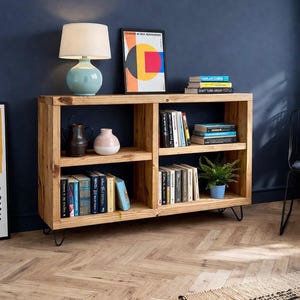 Op de afbeelding: Een lichtgekleurde houten boekenkast met meerdere planken, gevuld met boeken, decoratieve items en een kleine potplant. Een blauwe lamp en een ingelijst kunstwerk staan erop. De boekenkast staat op haarspeldpoten.