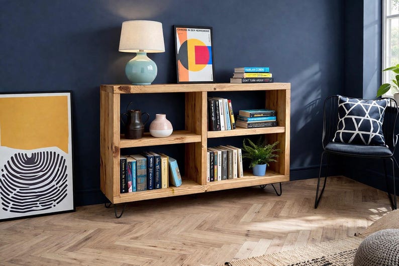 Op de afbeelding: Een lichtgekleurde houten boekenkast met meerdere planken, gevuld met boeken, decoratieve items en een kleine potplant. Een blauwe lamp en een ingelijst kunstwerk staan erop. De boekenkast staat op haarspeldpoten.