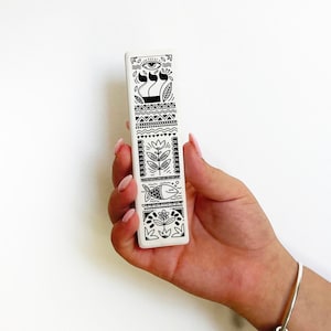 May include: A white ceramic item with black line art designs is held in a hand. The designs include an eye, flowers, fish, and geometric patterns. The item is approximately 15 cm long.