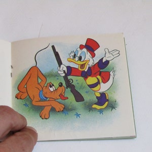 May include: A cartoon illustration of Donald Duck holding a rifle and pointing it at Pluto, a brown dog. Donald Duck is wearing a red and yellow outfit with a black and white striped pattern. Pluto is looking at Donald Duck with a scared expression. The background is a green field with blue flowers.