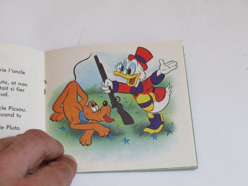 May include: A cartoon illustration of Donald Duck holding a rifle and pointing it at Pluto, a brown dog. Donald Duck is wearing a red and yellow outfit with a black and white striped pattern. Pluto is looking at Donald Duck with a scared expression. The background is a green field with blue flowers.