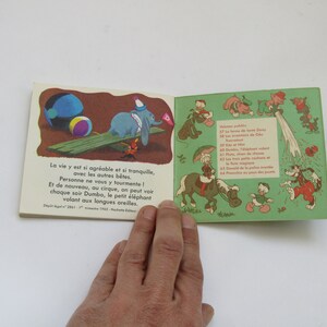 May include: Vintage children's book featuring illustrations of Dumbo the elephant and other Disney characters. The open pages display colorful drawings and French text. The book is held by a hand against a white background.