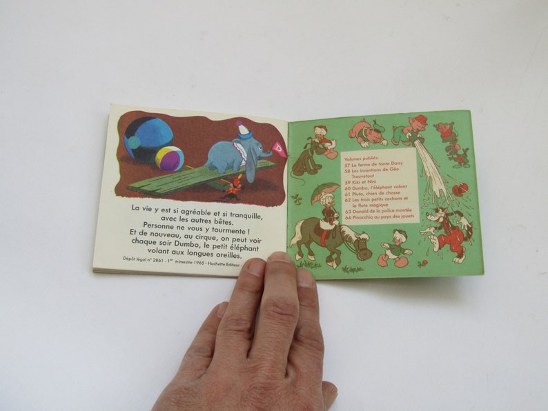 May include: Vintage children's book featuring illustrations of Dumbo the elephant and other Disney characters. The open pages display colorful drawings and French text. The book is held by a hand against a white background.