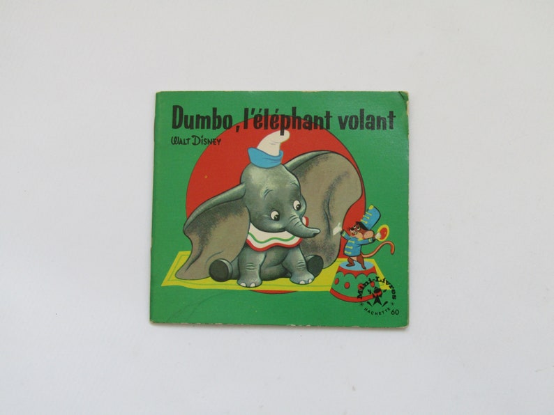May include: Vintage children's book cover featuring Dumbo the elephant. The cover is green with the title "Dumbo, l'&eacute;l&eacute;phant volant" and Walt Disney's name. Dumbo is depicted with a blue hat and a mouse in a circus outfit.