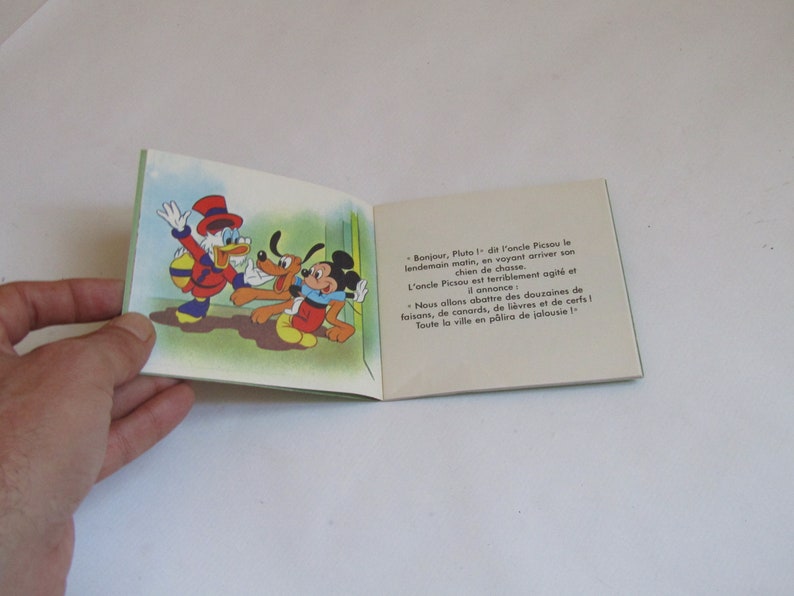 May include: A vintage children's book featuring Mickey Mouse, Pluto, and Donald Duck. The book's cover is green and features a cartoon illustration of the three characters. The text on the open page reads: "Bonjour, Pluto! -  de l'oncle Picsou le lendemain matin, en voyant arriver son chien de chasse. L'oncle Picsou est terriblement agit&eacute; et il annonce: Nous allons abattre des douzaines de faisans, de canards, de li&egrave;vres et de cerfs! Toute la ville en p&acirc;lira de jalousie!"