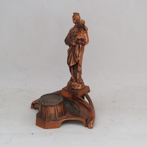 May include: A copper-colored statue of a saint holding a child, standing on a decorative base with three legs.
