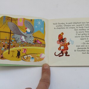 May include: Vintage children's book illustration featuring Dumbo the elephant flying in a circus. The image shows Dumbo with large ears, a circus tent, and a mouse character. Text in French is visible on the right page.