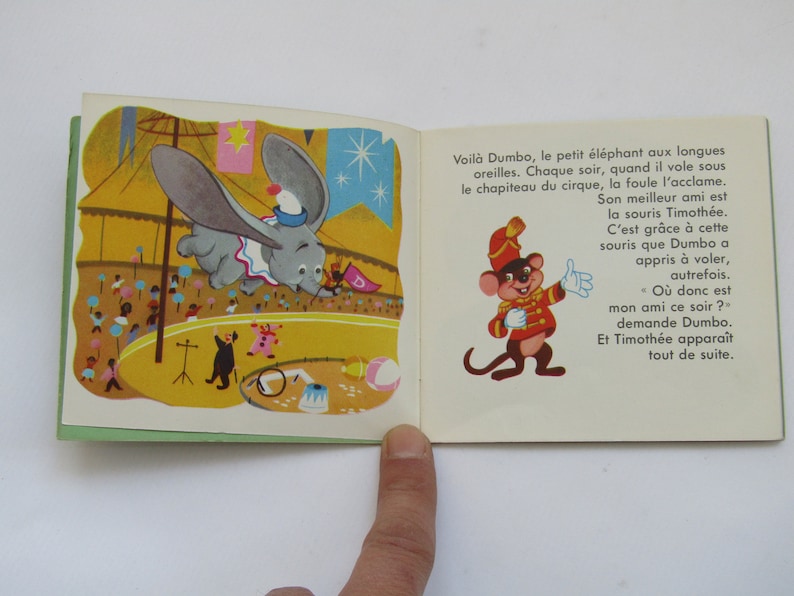May include: Vintage children's book illustration featuring Dumbo the elephant flying in a circus. The image shows Dumbo with large ears, a circus tent, and a mouse character. Text in French is visible on the right page.