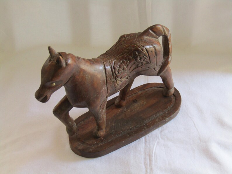 May include: A dark brown wooden horse sculpture with intricate carvings on its back and a detailed face. The horse is standing on a wooden base.