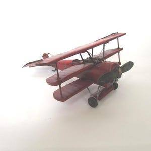 May include: A red metal triplane model featuring three stacked wings, a black propeller at the front, and small black wheels. The aircraft has a weathered finish with visible structural struts connecting the wings and a tail section at the rear.