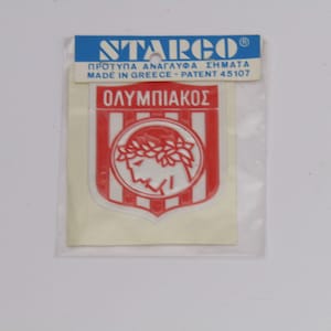 Peut inclure: Un autocollant en plastique rouge et blanc avec le logo de l'Olympiacos. Le logo représente un profil blanc d'un homme portant une couronne de laurier, sur un fond rayé rouge et blanc. Le texte "OLYMPIACOS" est imprimé en noir au-dessus du logo. L'autocollant est emballé dans un sac en plastique transparent avec une étiquette blanche qui indique "STARGO" et "MADE IN GREECE - PATENT 45107".