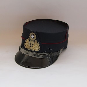 Könnte beinhalten: Eine dunkelblaue Militärmütze mit schwarzem Visier und goldenem Emblem. Die Mütze hat eine rote Paspel und einen Riemen. Das Emblem zeigt ein Wappen mit Kranz und Kreuz. Es handelt sich wahrscheinlich um einen Vintage- oder antiken Artikel.