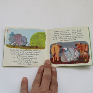 May include: Open book with colorful illustrations from a children's story. The left page shows Dumbo pulling a hay cart, while the right page depicts Dumbo with cows. Text in French accompanies the images.