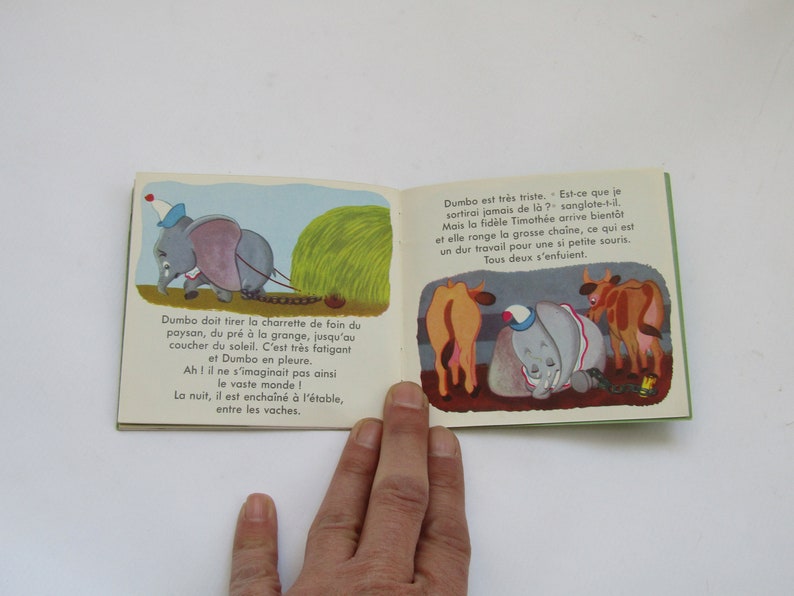 May include: Open book with colorful illustrations from a children's story. The left page shows Dumbo pulling a hay cart, while the right page depicts Dumbo with cows. Text in French accompanies the images.