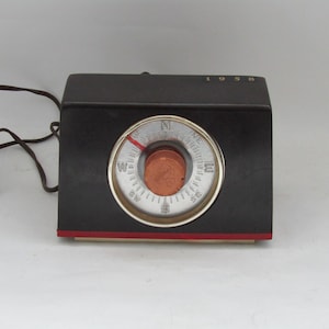 May include: A vintage compass in black and red with a silver face and a copper penny in the centre. The compass is labelled with the directions North, South, East, and West. The year 1958 is printed on the top of the compass.