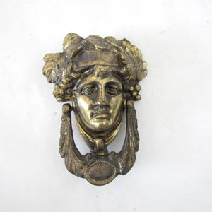 May include: Antique brass door knocker featuring a classical female face with detailed hair and a laurel wreath. The knocker has a ring-shaped handle and decorative leaf accents, adding a vintage touch to any door.