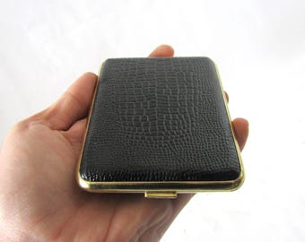 Könnte beinhalten: Ein rechteckiges schwarzes Etui mit einem strukturierten, krokodilartigen Muster. Das Etui hat eine goldfarbene Umrandung und wird in einer Hand vor weißem Hintergrund gehalten. Das Etui scheint geschlossen zu sein.