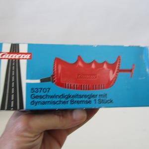 May include: A blue box with the Carrera logo and a red speed controller. The box has a black and white road graphic and the text "53707 Geschwindigkeitsregler mit dynamischer Bremse 1 Stück".