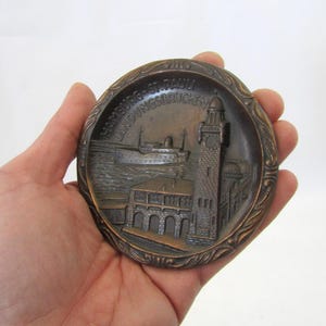 May include: A bronze-colored, round decorative plate featuring a detailed relief of a harbor scene with a lighthouse, buildings, and a ship. The plate has the text "HAMBURG ST. PAULI LANDUNGSBRUCKEN" around the edge.