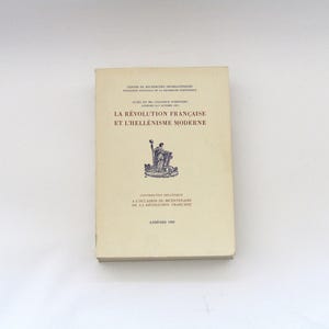 May include: A cream-coloured softcover book titled La Révolution Française et l'Hellénisme Moderne. The cover features dark red text, a central dark blue illustration of a figure holding a staff, and the publication location and year, Athènes 1989.