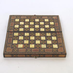 Könnte beinhalten: Ein rechteckiges, klappbares Spielbrett mit einem Raster aus abwechselnd cremefarbenen und dunklen Quadraten, die mit kleinen roten, grünen und schwarzen geometrischen Mustern verziert sind. Der Rand zeigt eine komplexe Holzeinlage.