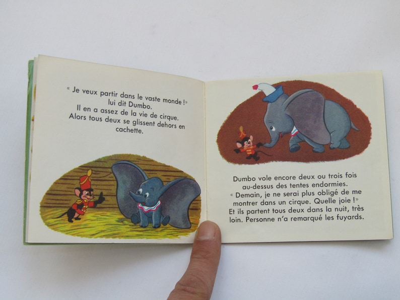 May include: Open book with illustrations from a children's story. The images depict Dumbo the elephant and a mouse in a circus setting. Text in French accompanies the illustrations.