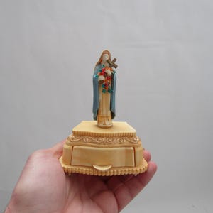 May include: A small, cream-colored, ornate box with a drawer. A figurine of a woman in a blue robe holding a cross and flowers is on top of the box.