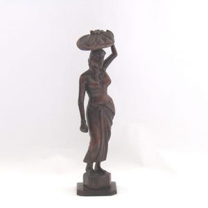 May include: A carved wooden statue of a woman carrying a basket on her head. The statue is brown and stands on a square base. The woman is draped in a long skirt and has one arm raised.