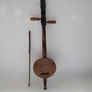 Op de afbeelding: Een houten muziekinstrument met een rond lichaam, een lange hals en een gesneden kop. Het instrument heeft twee stemknoppen en een enkele snaar. Het is een traditioneel instrument uit Zuidoost-Azië.