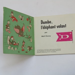 May include: Open vintage book titled "Dumbo, the Flying Elephant" by Walt Disney. The cover features illustrations of Dumbo and other Disney characters. The book is open to a page with the title and a pink flag with a white "D".