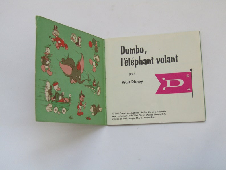 May include: Open vintage book titled "Dumbo, the Flying Elephant" by Walt Disney. The cover features illustrations of Dumbo and other Disney characters. The book is open to a page with the title and a pink flag with a white "D".